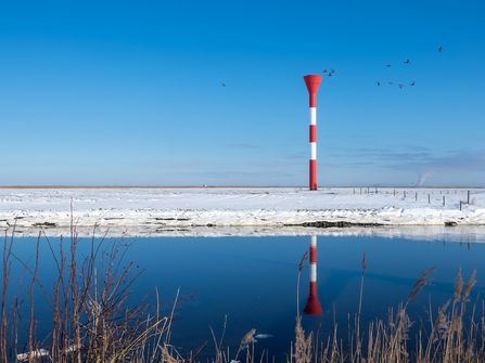 Das Bild zeigt das Otterndorfer Leuchtfeuer im Schnee im Hintergrund mit einem Teil der Hafeneinfahrt im Vordergrund.